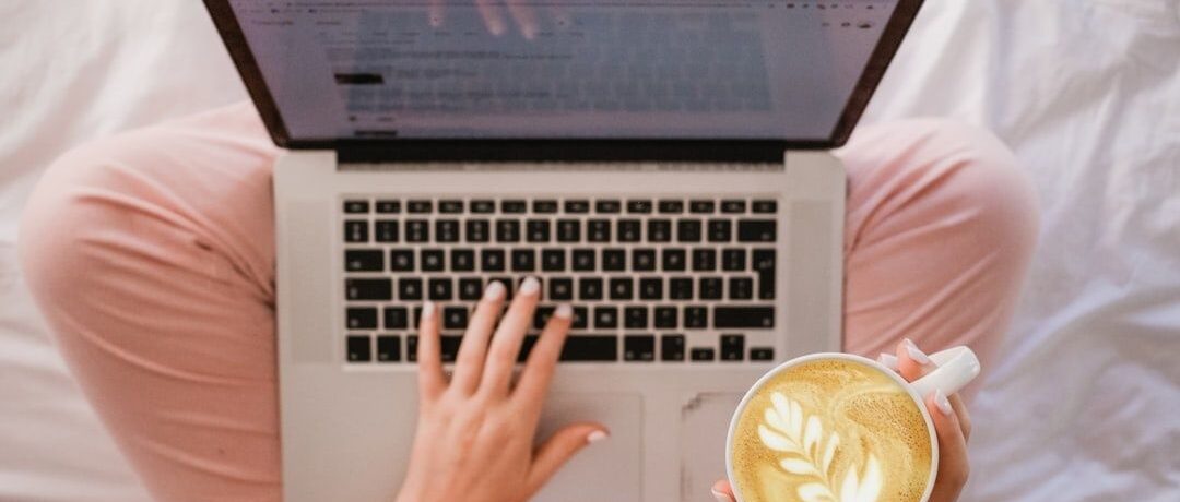 Person liest am Laptop im Bett und hält eine Tasse Kaffee