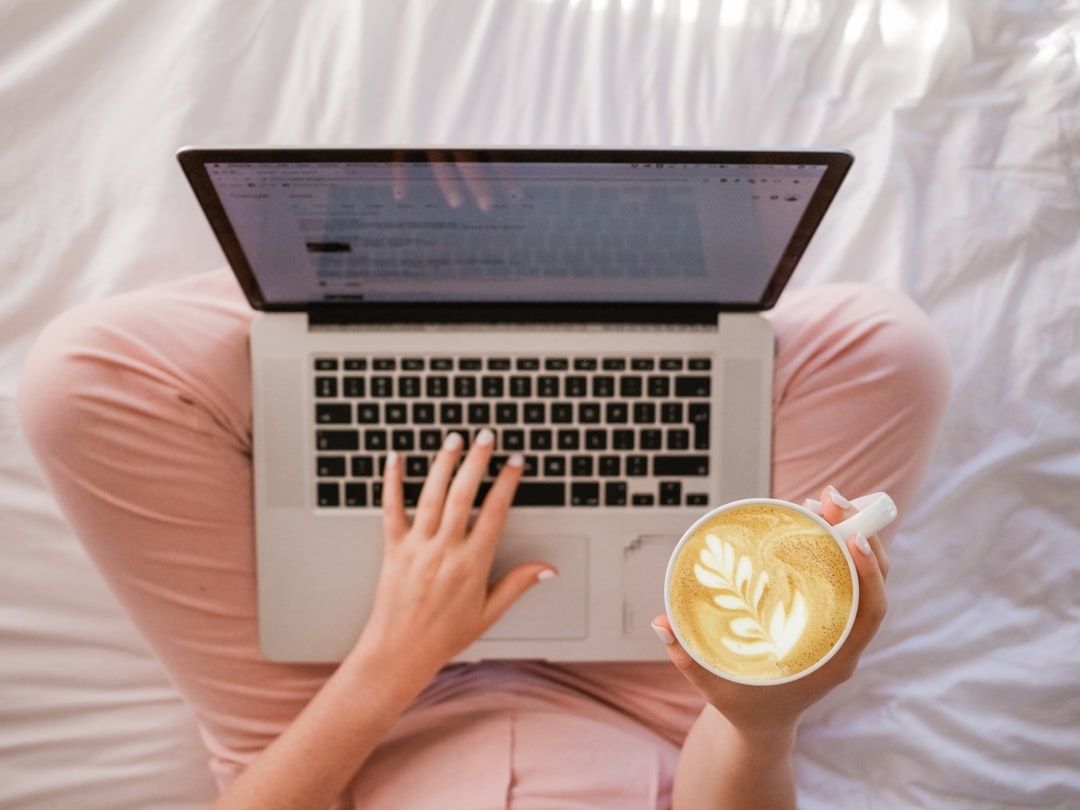 Person liest am Laptop im Bett und hält eine Tasse Kaffee